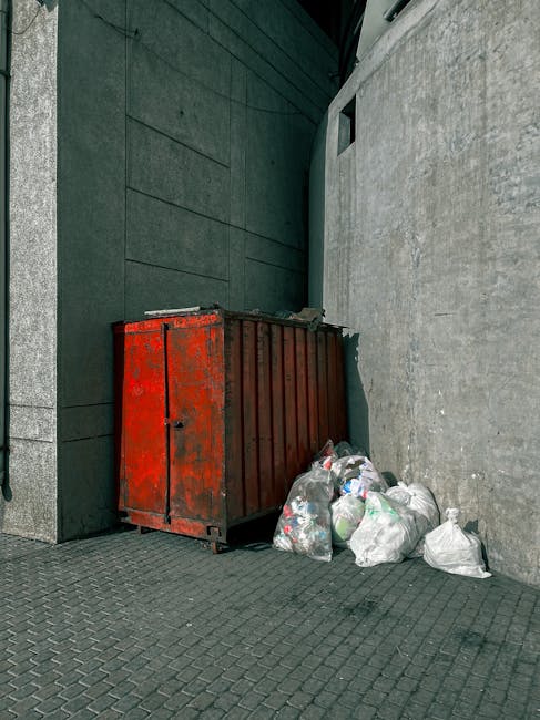 A red metal skip with a weathered finish is positioned against a grey concrete wall in an outdoor urban setting, likely a back alley or service area. The skip is rectangular, with visible vertical reinforcements on its sides and a slightly rusted surface indicating long-term outdoor use. Surrounding the base of the skip are several white plastic rubbish bags filled with mixed waste, some of which appear loosely tied and slightly bulging, suggesting they are hastily packed. The bags contain a variety of household waste and recyclables, as evidenced by the visible plastic bottles and packaging protruding from some of the bags. The pavement beneath is made of dark, textured concrete blocks that extend into the background, where a dark green wall or partition creates a narrow enclosure. On the right, part of a building’s exterior wall with a smooth, light grey finish is visible, along with a small, square window or vent high up on the wall. The scene is lit with natural daylight, with subtle shadows indicating an overcast sky or diffuse lighting conditions. The image reflects a typical scene for private waste collection or rubbish removal services in an urban environment, supported by companies like rubbishremovaledgware.com for alternative disposal methods outside council collection. During rubbish removal, such arrangements are common for curbside or back alley clearance, highlighting the importance of efficient waste management solutions in densely built areas.