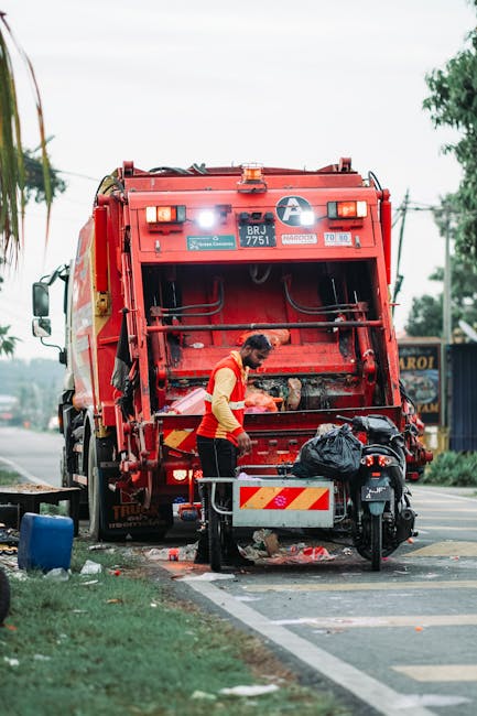 A street scene featuring a red waste collection vehicle, with the back compartment open for rubbish loading, parked beside a curb on a roadway. A sanitation worker, dressed in a red and yellow high-visibility vest and black trousers, is seen emptying a black plastic rubbish bag into the vehicle's rear compartment. The worker is focused on the task, with various debris and discarded waste visible on the ground near their feet. Adjacent to the waste truck, a black motorbike is parked with a small storage box attached to its side, which appears to contain additional rubbish or tools. In the background, there are trees, utility poles with overhead wires, and a distant structure, indicating an urban or suburban environment. The overall lighting suggests a daytime setting with cloudy or overcast skies. This scene exemplifies on-site waste collection as an alternative to traditional bin collection, highlighting the service provided by companies like rubbishremovaledgware.com for private rubbish removal or relocation without reliance on local authority services.
