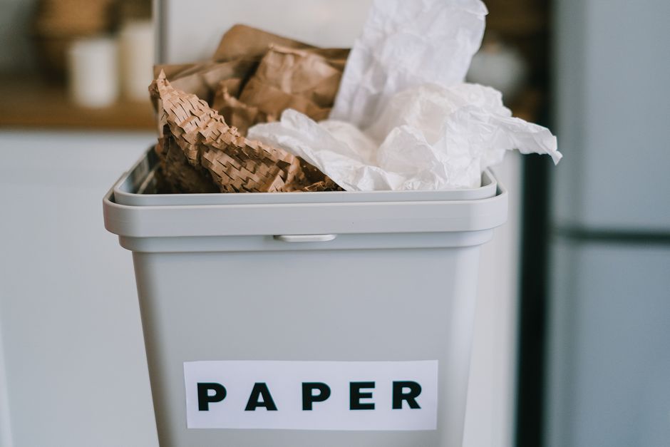A light grey rectangular paper recycling bin placed against a neutral background, filled with crumpled and torn sheets of white and brown paper, with some torn pieces and waste paper spilling over the top edge. The bin has a removable lid with a small integrated handle, and a white label with bold black letters spelling 'PAPER' adhered to the front. The surrounding environment appears to be a domestic or office setting, with a glimpse of furniture or appliances in the background, emphasizing the use of a dedicated container for paper waste collection in compliance with waste management guidelines. The lighting is natural, highlighting the textures of the crumpled paper and the smooth finish of the bin, supporting the context of responsible rubbish separation and informal disposal practices aligned with independent waste handling services in Edgware.