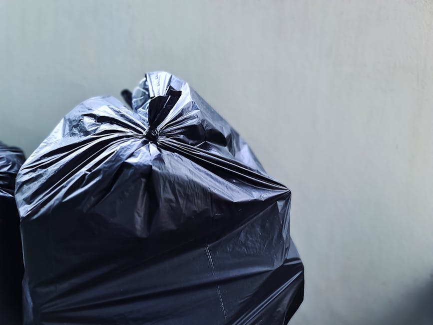 A close-up view of several black plastic rubbish bags filled with waste, stacked against a light-colored, smooth wall. The bags are tightly knotted at the top, with some showing creases and reflections on the shiny plastic surface. The bags are placed on a flat, possibly paved or concrete surface, suggesting an outdoor or industrial environment suitable for private waste disposal or collection services. No other objects or environmental features are visible, emphasizing the focus on the waste containment. The scene conveys a typical scene for independent rubbish collection in commercial or retail areas, with the rubbish ready for pickup or disposal by a service such as Rubbish Removal Edgware. The lighting appears natural, and the image's sharpness highlights the textures and details of the plastic bags.