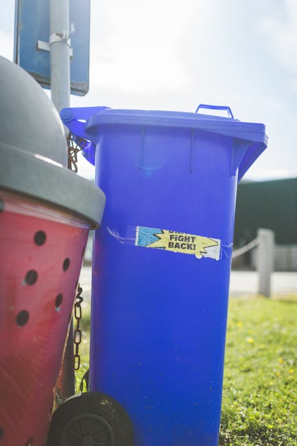A large collection of overflowing rubbish bins and mixed waste scattered around a designated collection point in an urban setting, situated on a paved sidewalk adjacent to a parking area. The scene includes a grey mixed paper and card waste bin with an open lid, overflowing with flattened cardboard boxes, crumpled paper, and packaging materials. Surrounding the bin are black and red rubbish bags, some partially open revealing used packaging, along with various cardboard boxes, plastic waste, and loose paper strewn across the pavement. To the left, a parked grey car is positioned behind a metal railing, with a tree nearby. The background features a commercial building with blue scaffolding covering the upper part, indicating ongoing maintenance or renovation, and a row of storefronts with signs, including a fish bar, visible under large windows. The scene suggests an instance of unmanaged or neglected waste typical of an independent rubbish collection service, highlighting the importance of regular rubbish removal for shop fronts and public spaces.