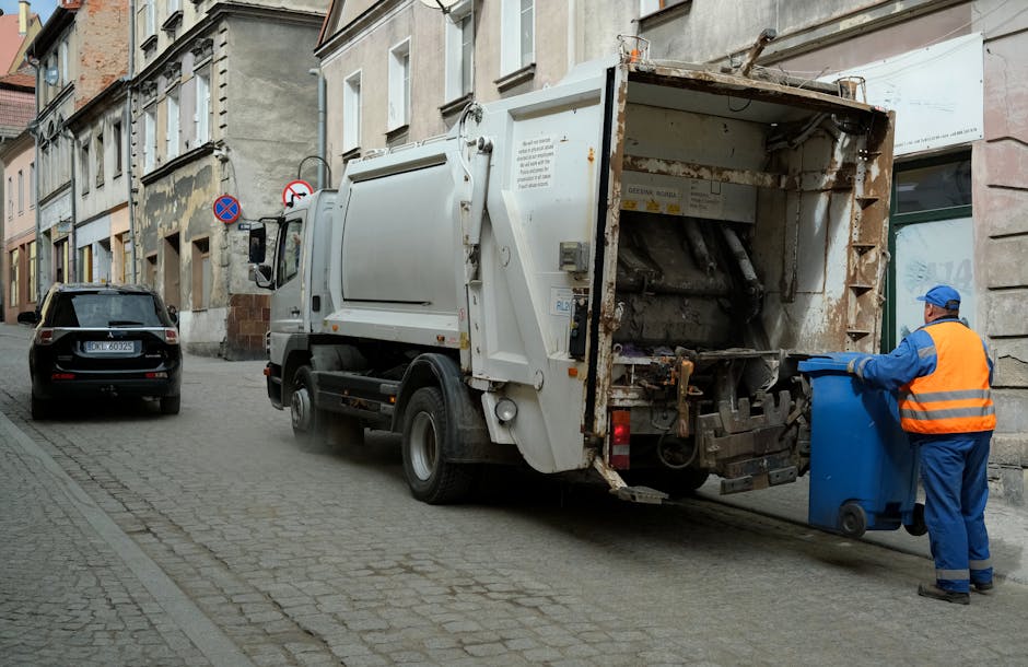 A sanitation worker wearing a blue uniform, orange high-visibility vest, and a blue cap is standing on the cobblestone street next to a large white rubbish collection vehicle, actively emptying a blue wheelie bin into the rear loading compartment. The rubbish truck, which shows signs of rust and dirt on its metal surfaces, is positioned adjacent to a multi-story building with a weathered facade, featuring a mix of brick and plaster walls. The vehicle's rear section is open, revealing mechanical components used for compacting waste. In the background, there are parked cars, including a black vehicle with a visible license plate, and street signs indicating parking restrictions and no stopping zones. The environment appears to be part of an urban residential area with narrow streets and a slightly overcast sky. The scene is consistent with on-site waste collection or private rubbish removal services typical of independent waste disposal operations coordinated by companies like rubbishremovaledgware.com.